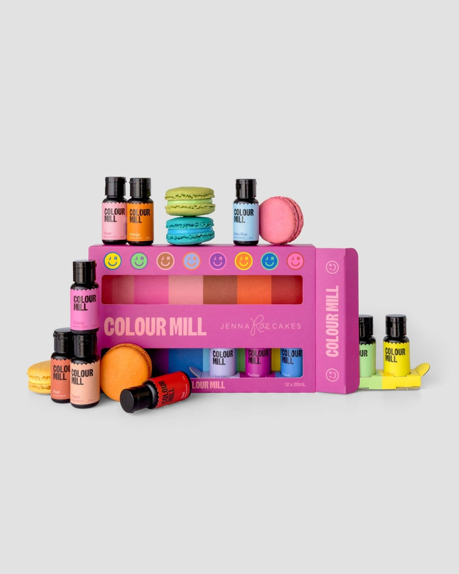 The Jenna Rae Cakes x Colour Mill Pack by Jenna Rae Cakes features multiple small food coloring bottles arranged in and around a pink box, displayed with colorful macarons and fondant shapes on a light gray background.