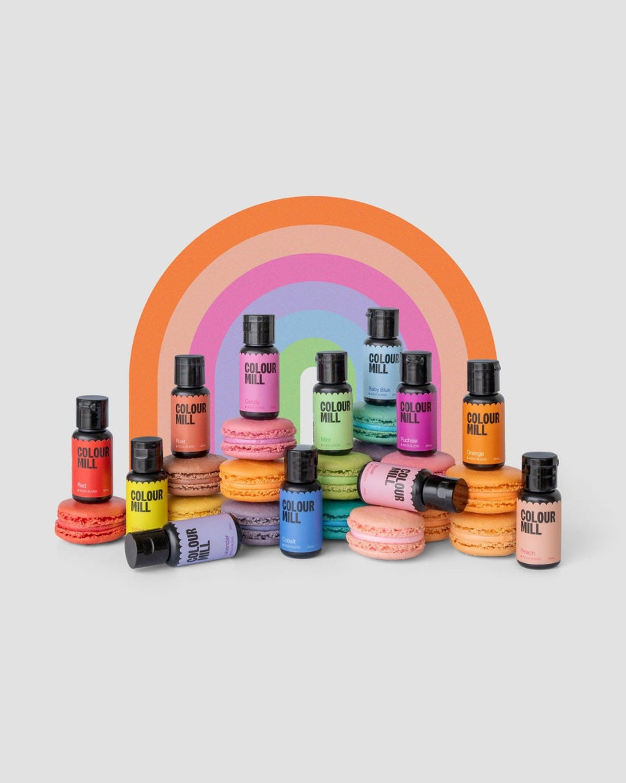 Jenna Rae Cakes x Colour Mill Pack bottles are displayed with pastel macarons in front of a vibrant rainbow arch, all set against a light grey background.