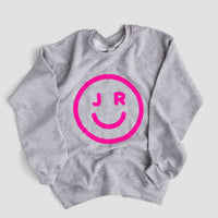 The JRC Smiley Sweatshirt by Jenna Rae Cakes is a gray top with a bold pink smiley face and the letters J and R as eyes printed on the front.