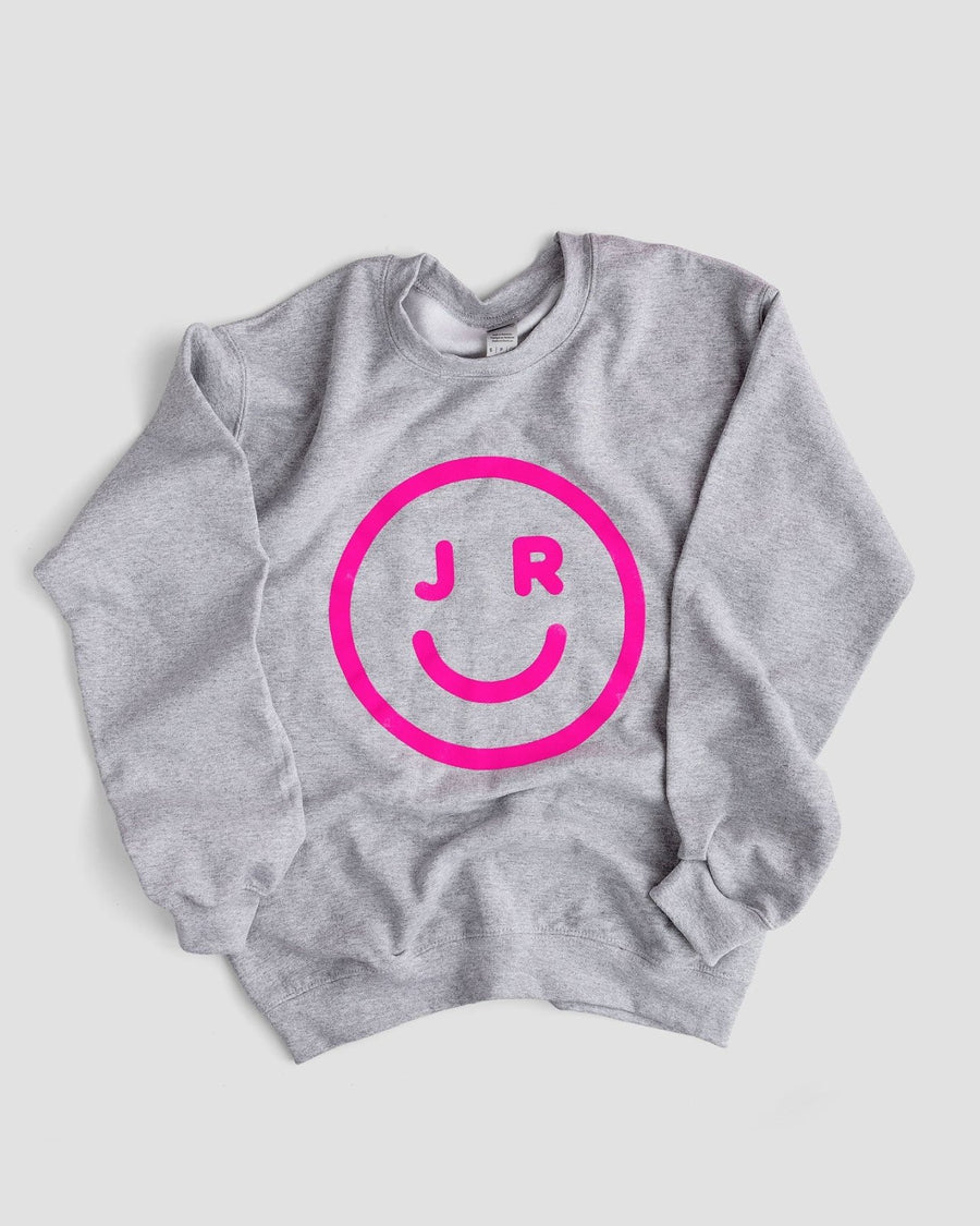 The JRC Smiley Sweatshirt by Jenna Rae Cakes is a gray top with a bold pink smiley face and the letters J and R as eyes printed on the front.