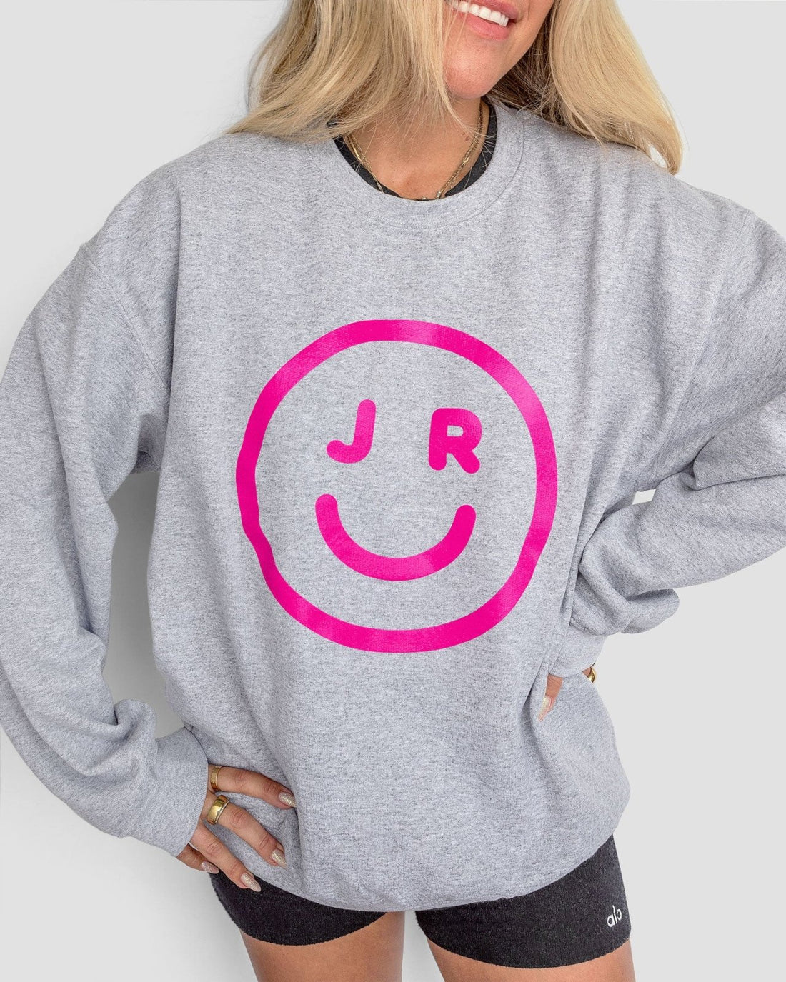 A person wearing the Jenna Rae Cakes JRC Smiley Sweatshirt—a light gray top featuring a large pink smiley face with J R inside—paired with black shorts. Their blonde hair is down, and theyre smiling with one hand on their hip.