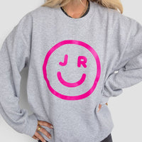A person wearing the Jenna Rae Cakes JRC Smiley Sweatshirt—a light gray top featuring a large pink smiley face with J R inside—paired with black shorts. Their blonde hair is down, and theyre smiling with one hand on their hip.
