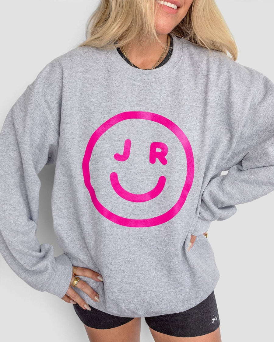 A person wearing the Jenna Rae Cakes JRC Smiley Sweatshirt—a light gray top featuring a large pink smiley face with J R inside—paired with black shorts. Their blonde hair is down, and theyre smiling with one hand on their hip.