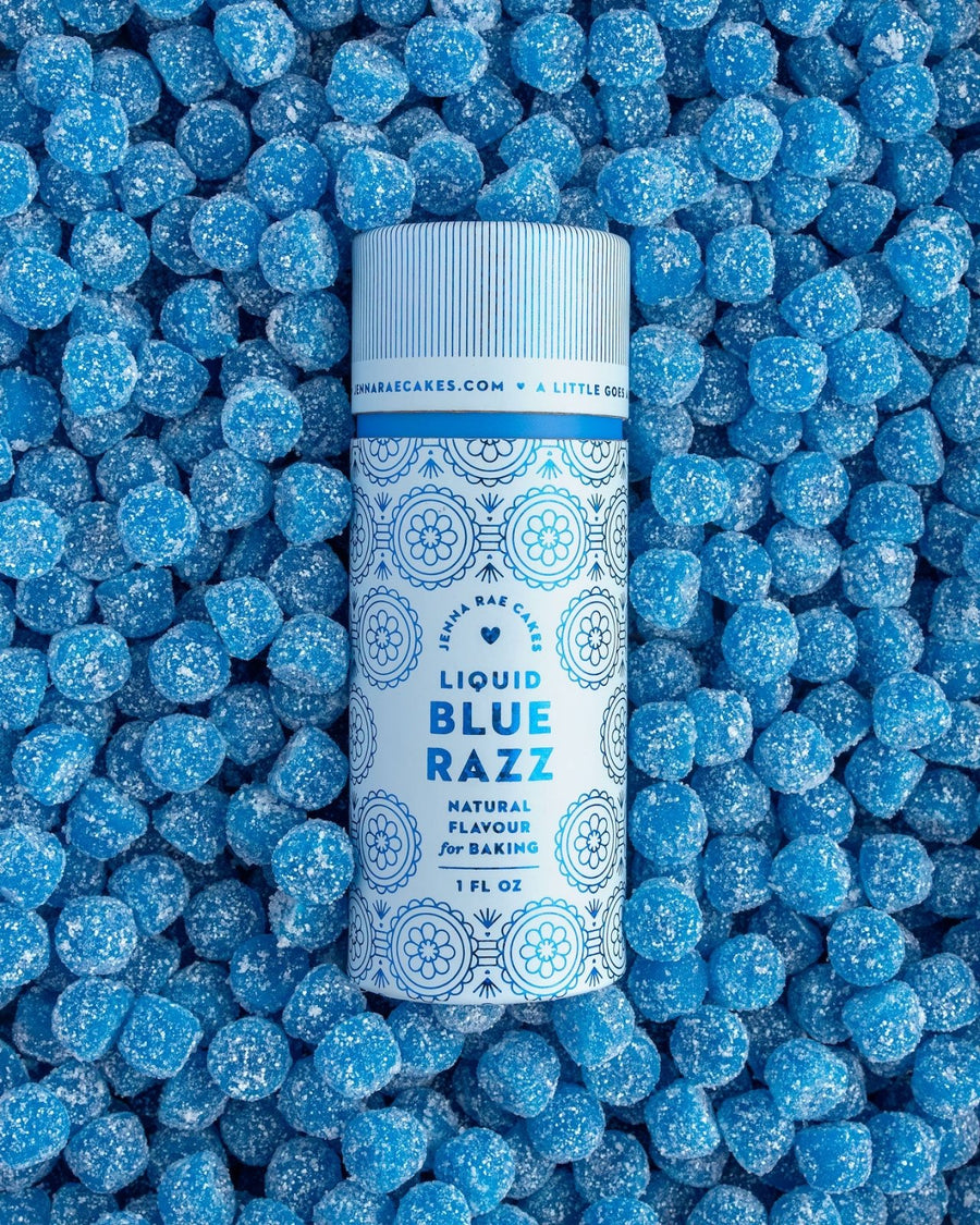 A bottle of Jenna Rae Cakes Liquid Blue Razz stands upright on a layer of small, round, blue sugar-coated candies, creating a vibrant blue-themed background.