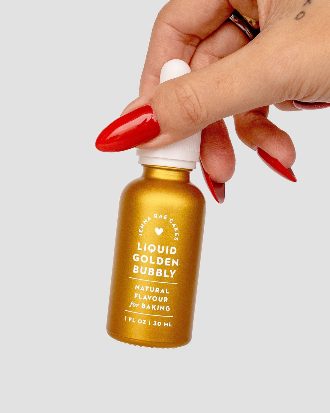 A hand with long red nails holds a small gold bottle of Jenna Rae Cakes Liquid Golden Bubbly against a plain light background.