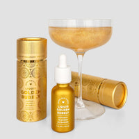 A coupe glass of sparkling golden liquid sits behind a dropper bottle and two cylindrical containers of Jenna Rae Cakes’ Liquid Golden Bubbly Natural Flavor for Baking in shiny gold packaging.