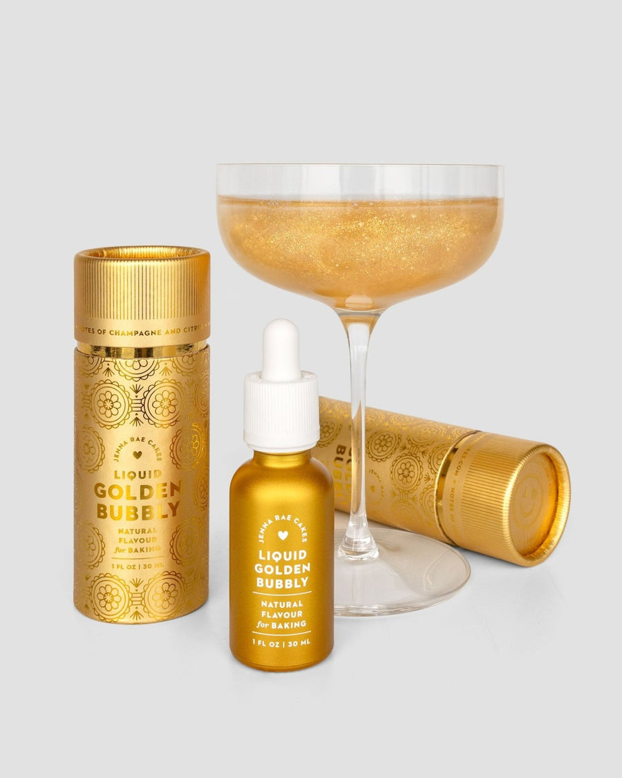 A champagne coupe of sparkling gold sits behind a gold dropper bottle and two gold cylinders labeled Liquid Golden Bubbly - Package of 6 by Jenna Rae Cakes, a natural flavour perfect for baking.