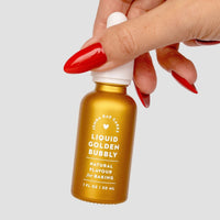 A hand with long red nails holds a small gold bottle of Jenna Rae Cakes Liquid Golden Bubbly - Package of 6, Natural Flavour for Baking, 1 FL OZ / 30 mL, against a plain light background.