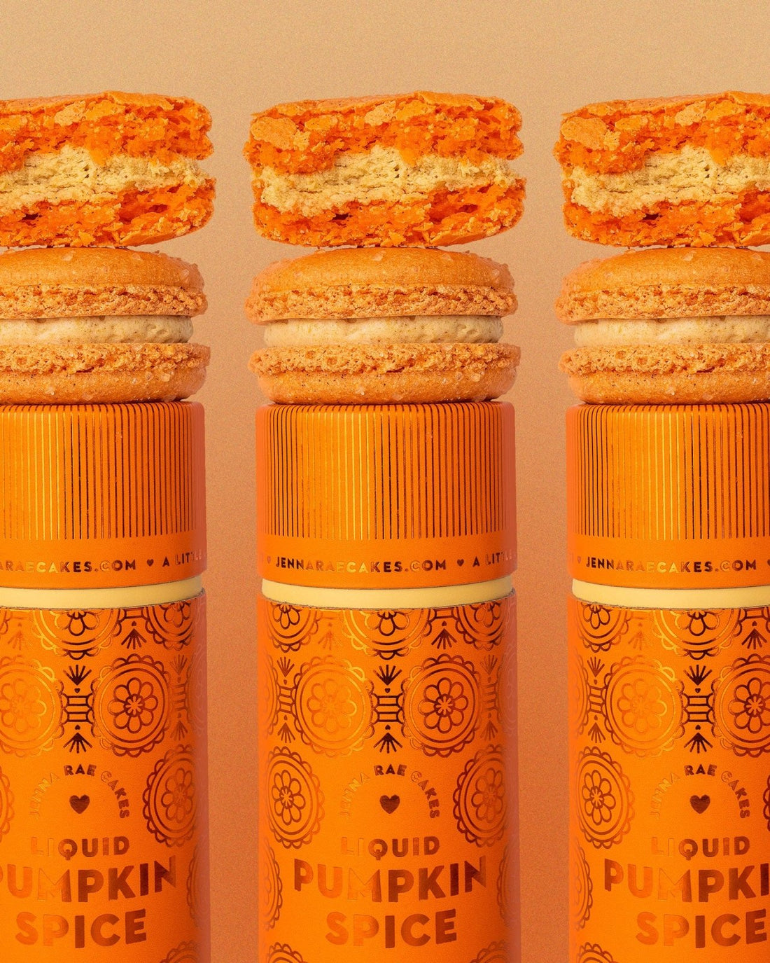 Three orange Jenna Rae Cakes Liquid Pumpkin Spice canisters are each topped with a pumpkin macaron and pumpkin-flavored cookie, featuring decorative patterns and branding on a light background.