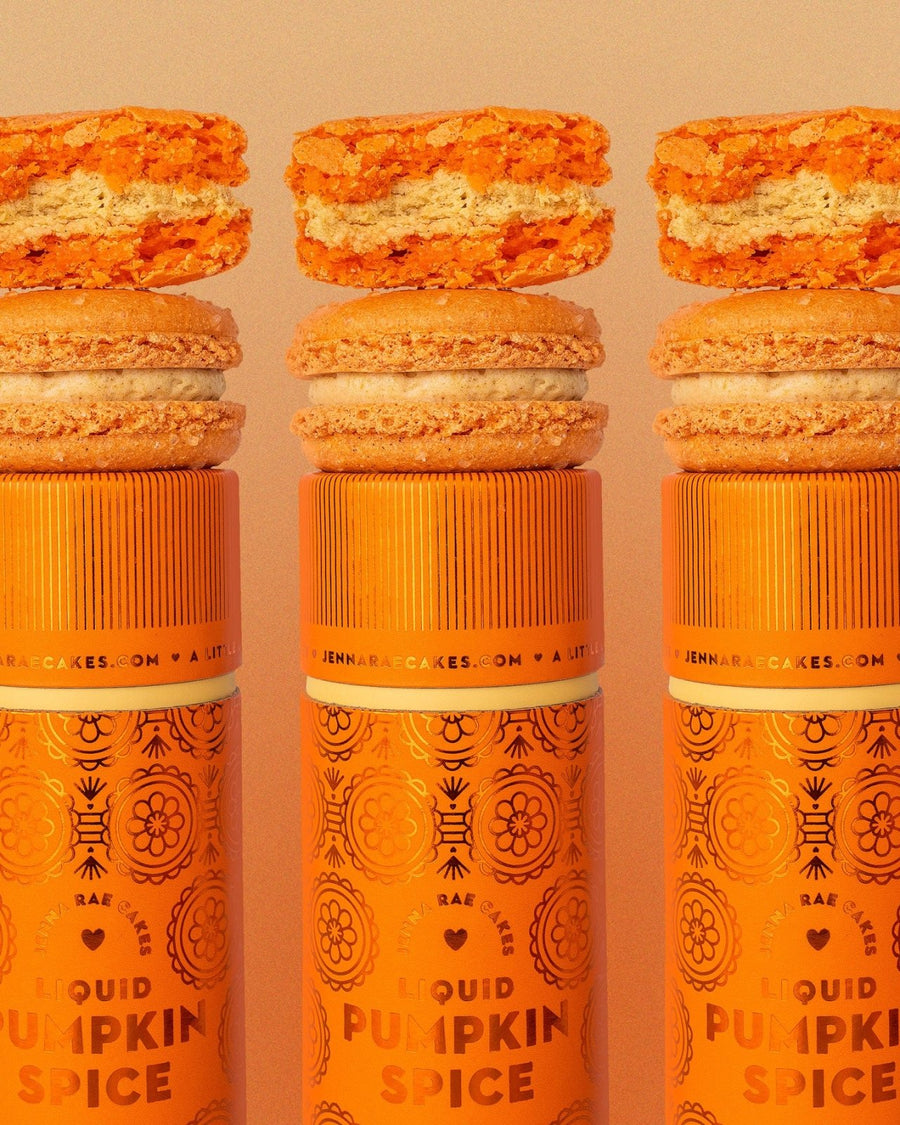 Three orange Jenna Rae Cakes Liquid Pumpkin Spice canisters are each topped with a pumpkin macaron and pumpkin-flavored cookie, featuring decorative patterns and branding on a light background.