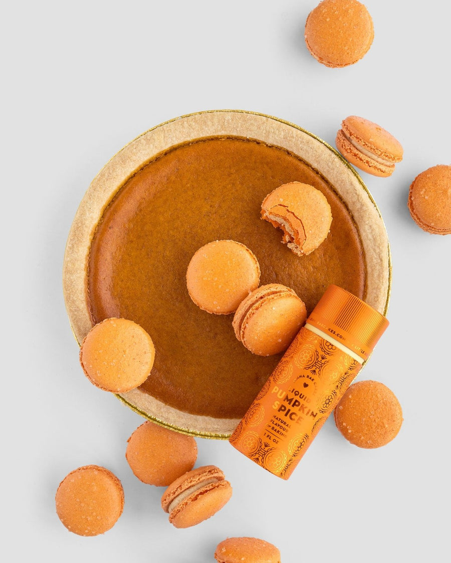 A pumpkin pie surrounded by orange macarons, one partially broken, is shown with a Jenna Rae Cakes Liquid Pumpkin Spice container on a light gray background.