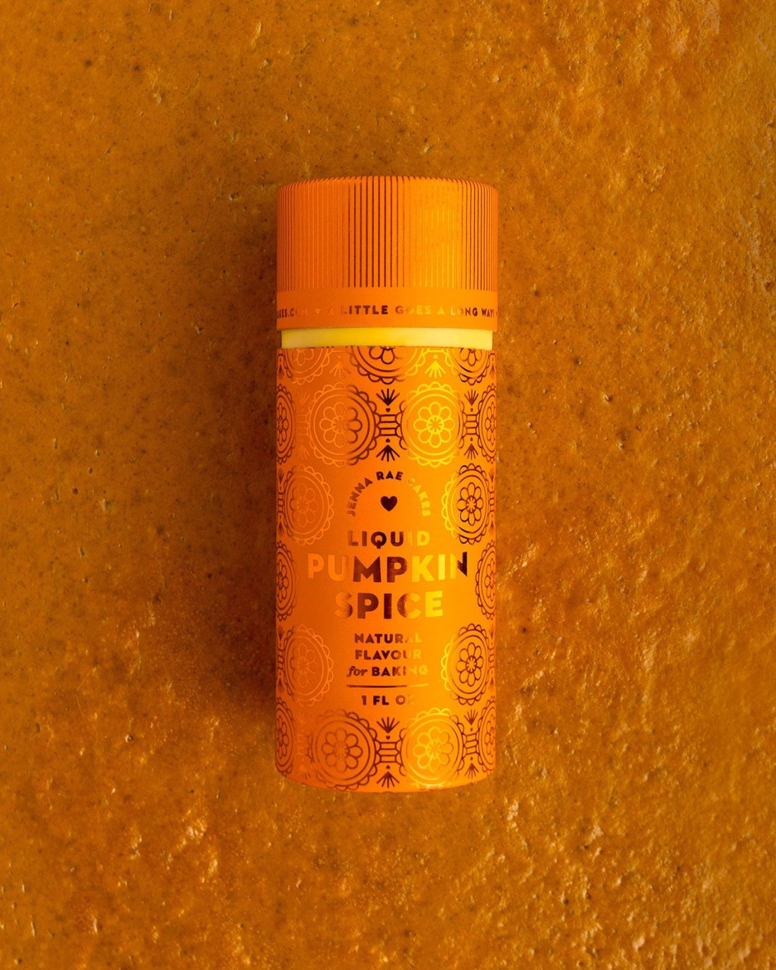 A small orange bottle of Jenna Rae Cakes Liquid Pumpkin Spice rests on an orange textured surface, adorned with decorative patterns and labeled as a natural baking flavor.