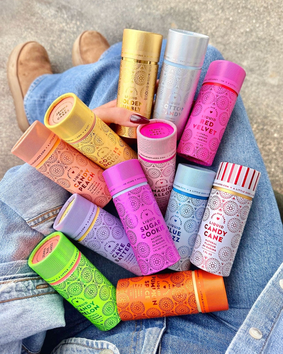 A hand holds an assortment of colorful, patterned bottles labeled with flavors like Golden Toffee Caramel, Red Velvet, Sugar Cookie, and Candy Cane, resting on denim-clad legs and tan shoes.