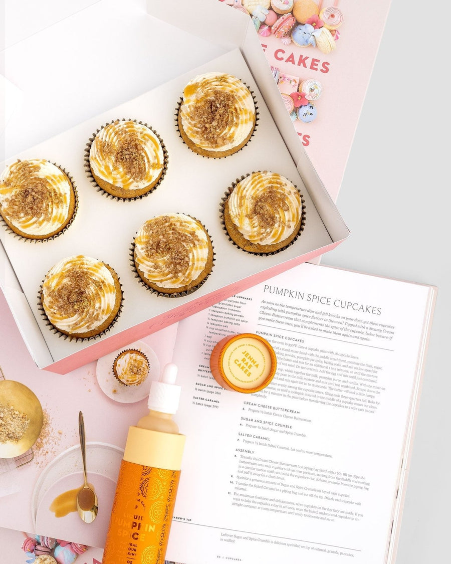 A package of six Jenna Rae Cakes Liquid Pumpkin Spice sits next to an open cookbook with a pumpkin spice cupcake recipe, along with a frosting bottle, small jar, spoon, and colorful sprinkles scattered on the table.