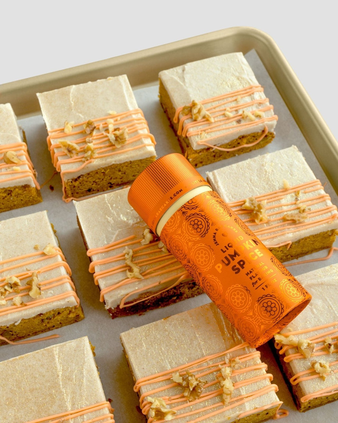A baking tray with frosted pumpkin spice bars, topped with orange icing and nuts, is shown. A Jenna Rae Cakes Liquid Pumpkin Spice - Package of 6 bottle rests diagonally on the bars.