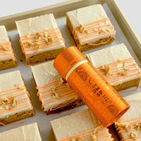 A baking tray with frosted pumpkin spice bars, topped with orange icing and nuts, is shown. A Jenna Rae Cakes Liquid Pumpkin Spice - Package of 6 bottle rests diagonally on the bars.