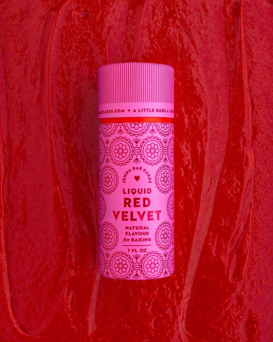 A pink bottle of Jenna Rae Cakes Liquid Red Velvet stands upright against a textured red background with swirls. The packaging features circular decorative designs and bold red lettering.