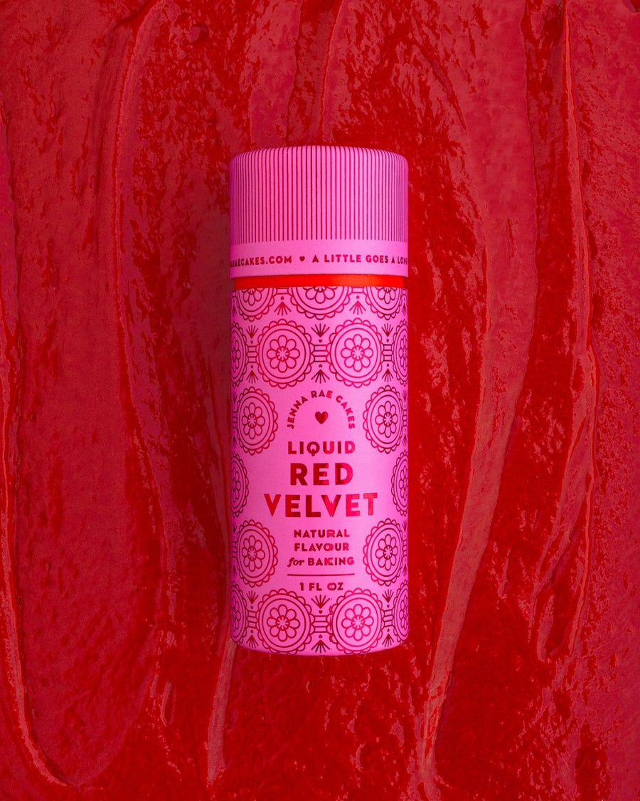 A pink bottle of Jenna Rae Cakes Liquid Red Velvet stands upright against a textured red background with swirls. The packaging features circular decorative designs and bold red lettering.