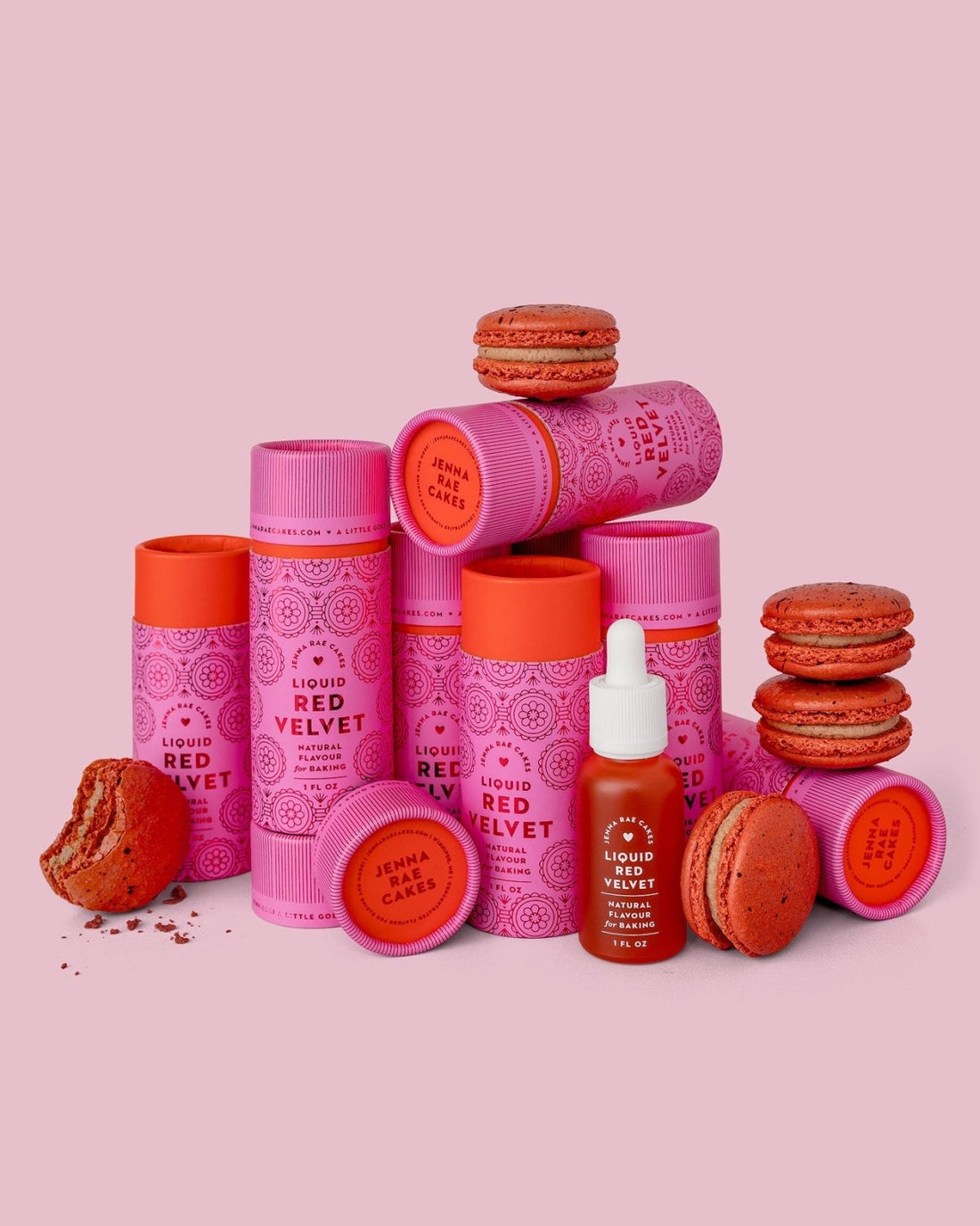 Jenna Rae Cakes’ Liquid Red Velvet - Package of 6 is displayed in pink and red cylindrical containers alongside stacked and broken red macarons and a small dropper bottle, all set against a pink background.