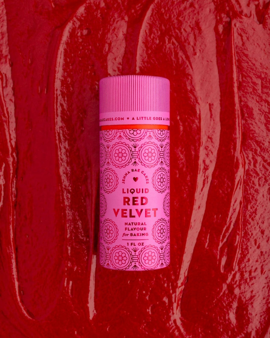 A package of 6 Jenna Rae Cakes Liquid Red Velvet bottles sits on a textured red surface, resembling cake batter or frosting, with decorative patterns and text on the pink labels.