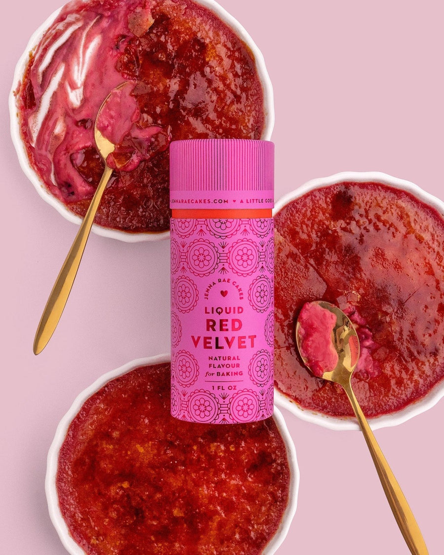 Three bowls of red velvet crème brûlée are topped with caramelized sugar and gold spoons. In the center sits a pink Jenna Rae Cakes Liquid Red Velvet - Package of 6, perfect for baking and drinks.