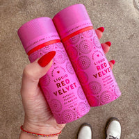 A hand with red-painted nails holds two cans of Jenna Rae Cakes Liquid Red Velvet (Package of 6) against a concrete background. The person wears white shoes and beaded bracelets.