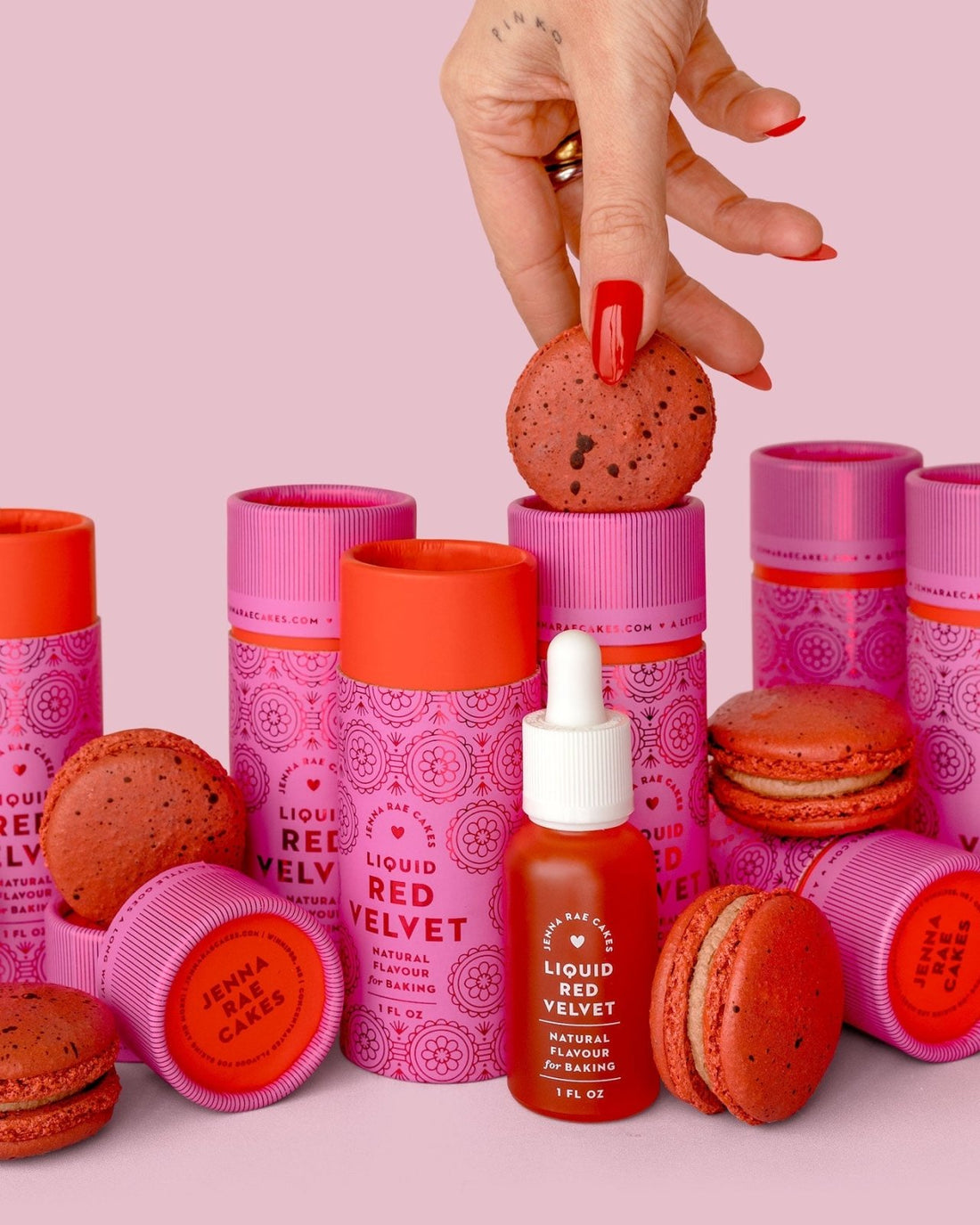A hand with red nails holds a red macaron above a set of pink and red Jenna Rae Cakes Liquid Red Velvet - Package of 6 bottles and matching macarons, all arranged on a pink background.
