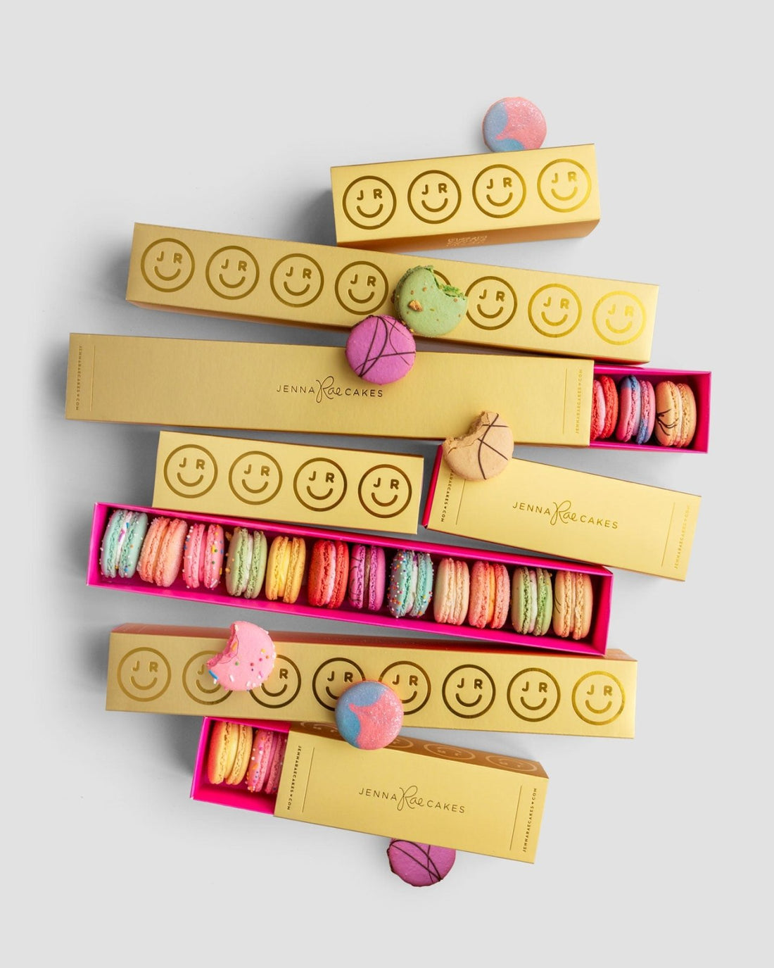 Jenna Rae Cakes Macaron Gift Box features long, rectangular gold boxes filled with colorful macarons, some of which are scattered around the boxes on a light gray background.