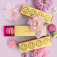 Three Jenna Rae Cakes Macaron Gift Boxes are surrounded by pink and purple flowers, with pink, green, and sprinkled macarons scattered on a light pink background.