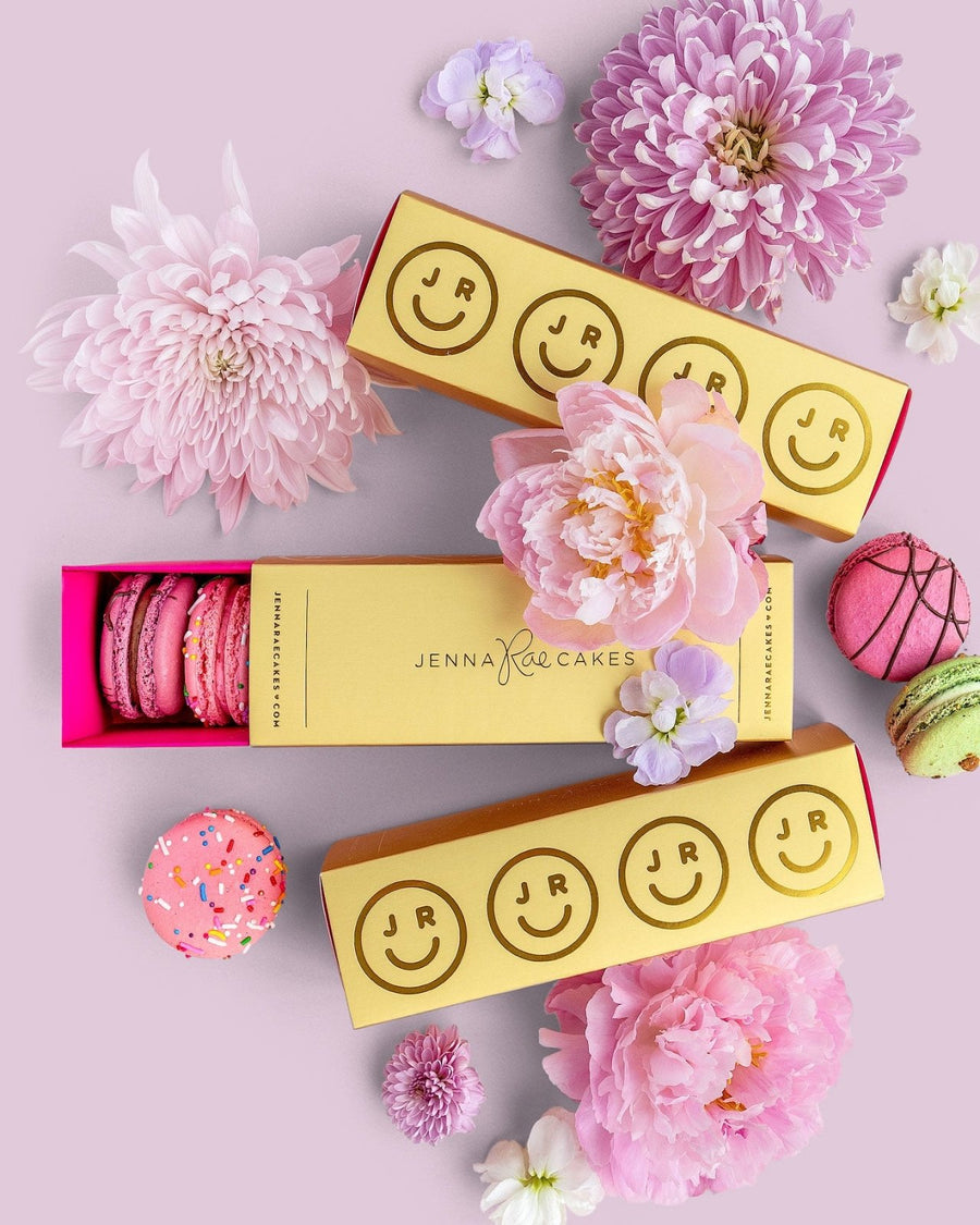 Three Jenna Rae Cakes Macaron Gift Boxes are surrounded by pink and purple flowers, with pink, green, and sprinkled macarons scattered on a light pink background.