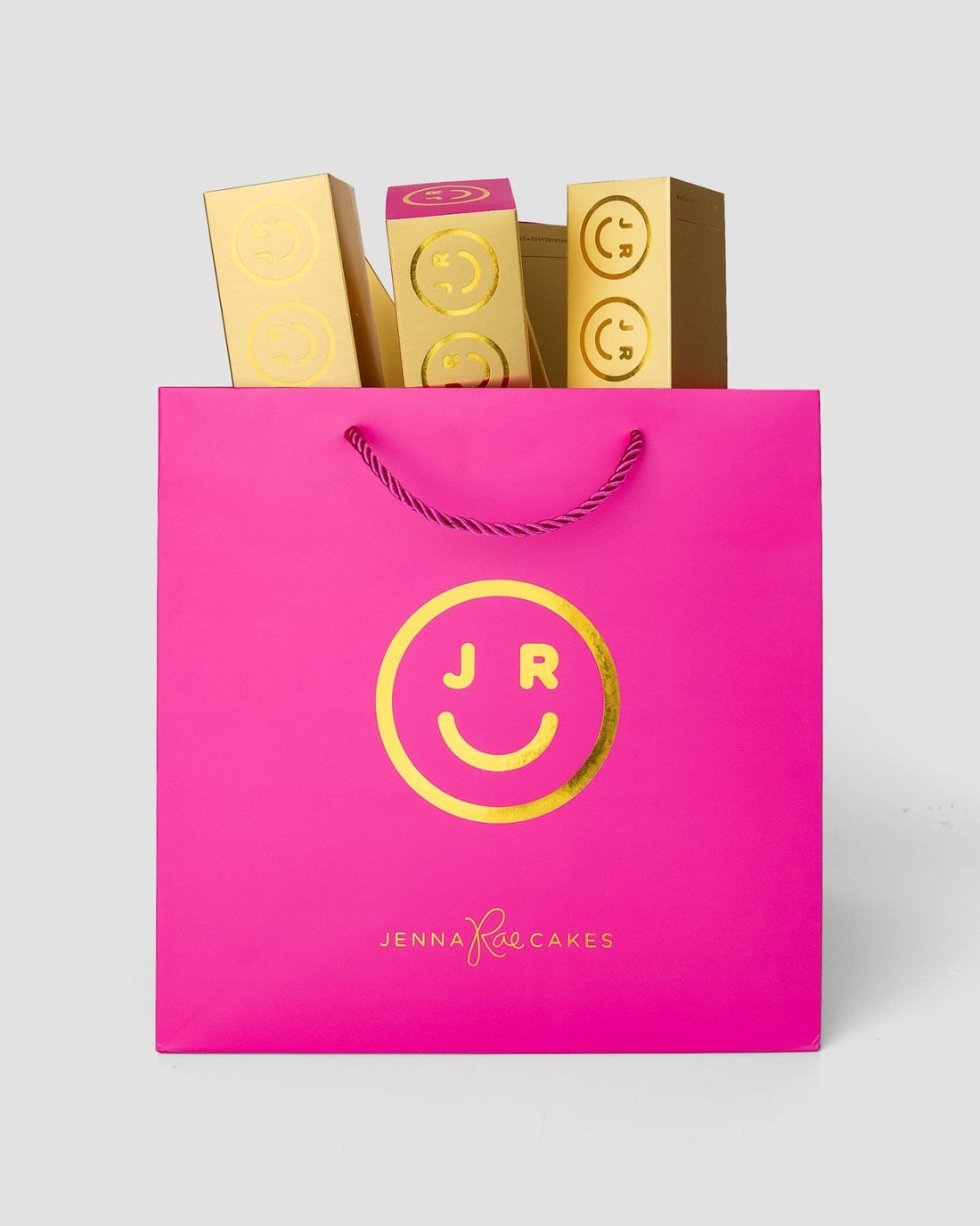A pink Jenna Rae Cakes shopping bag with a gold smiley face, holding Macaron Gift Boxes, each decorated with matching gold smiley faces and JR letters.