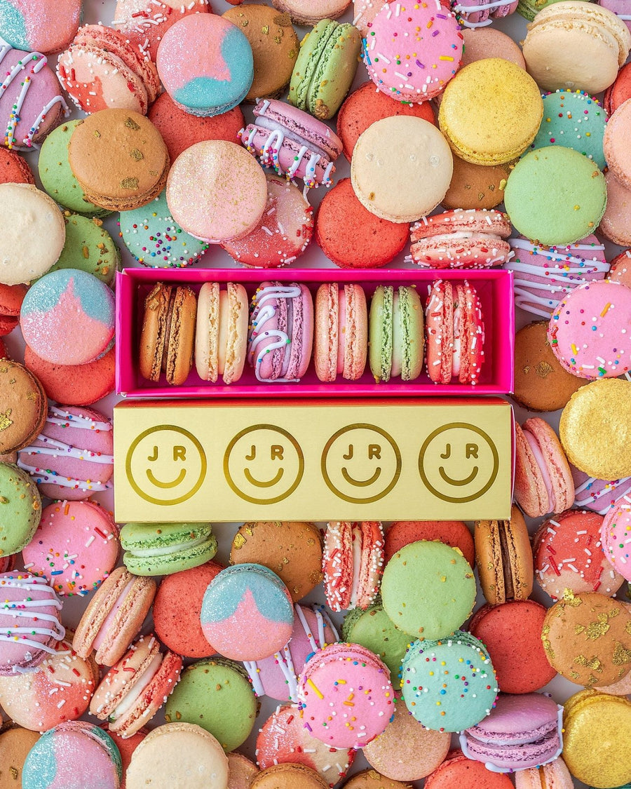 An open yellow Jenna Rae Cakes Macaron Gift Box with a pink interior displays six colorful, sprinkle- and icing-decorated macarons. The box features smiley faces and JR branding, surrounded by assorted macarons.