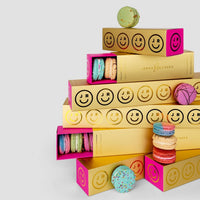 A stack of Jenna Rae Cakes Macaron Gift Boxes, adorned with smiley faces, is surrounded by colorful macarons against a plain light gray background.