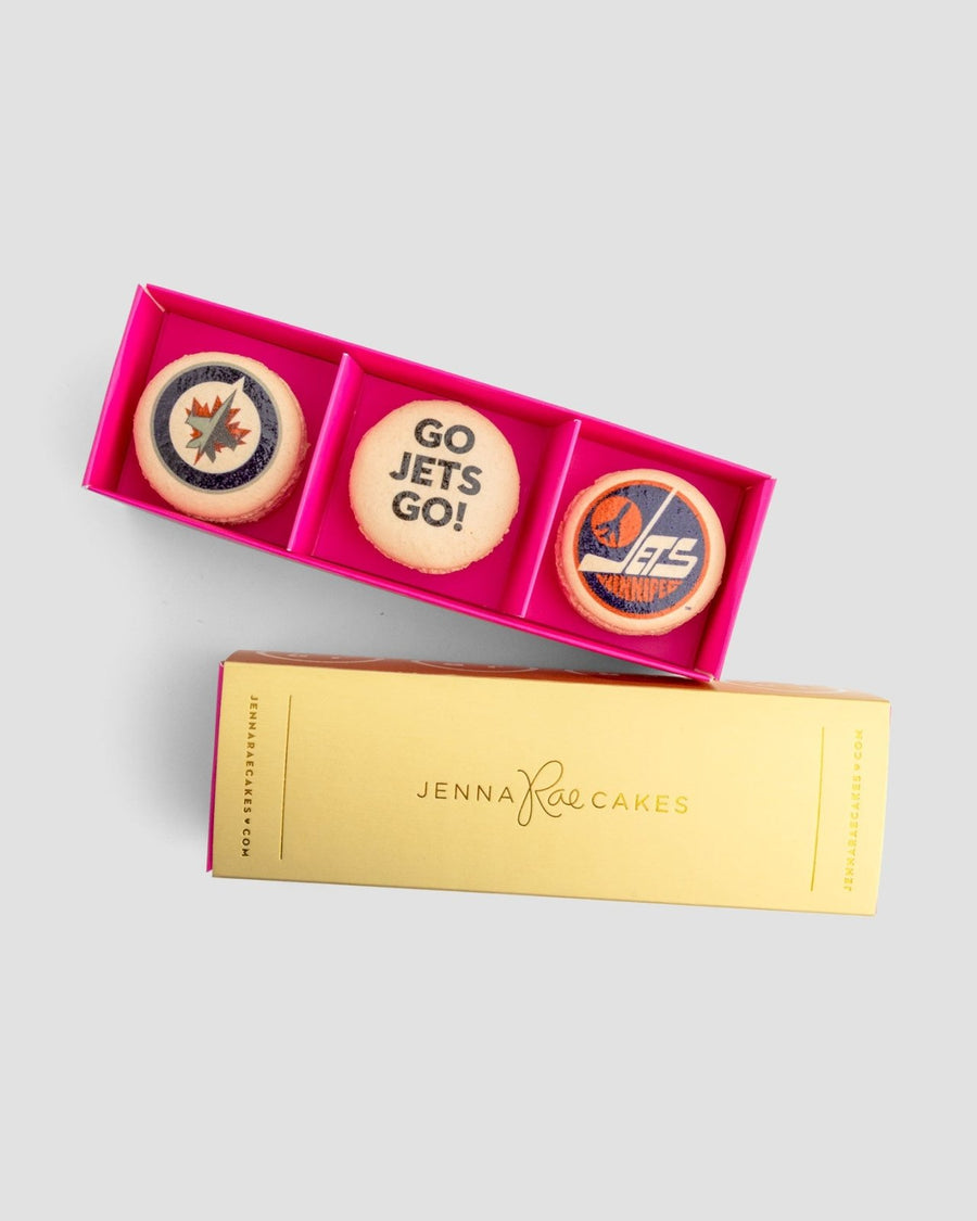 The Macaron Message Gift Box by Jenna Rae Cakes features three macarons in a pink box—two with Winnipeg Jets logos and one with GO JETS GO!—displayed beside a yellow Jenna Rae Cakes sleeve.