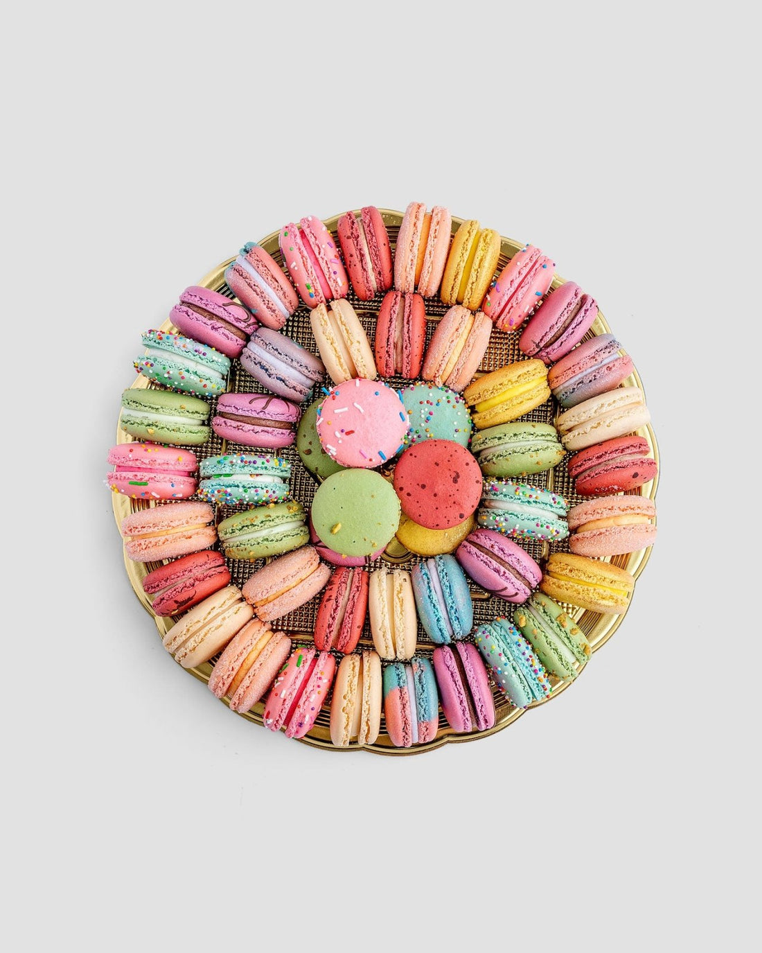 The Jenna Rae Cakes Macaron Party Platter features colorful macarons arranged in concentric circles around round cookies at the center, all beautifully displayed on a white background.
