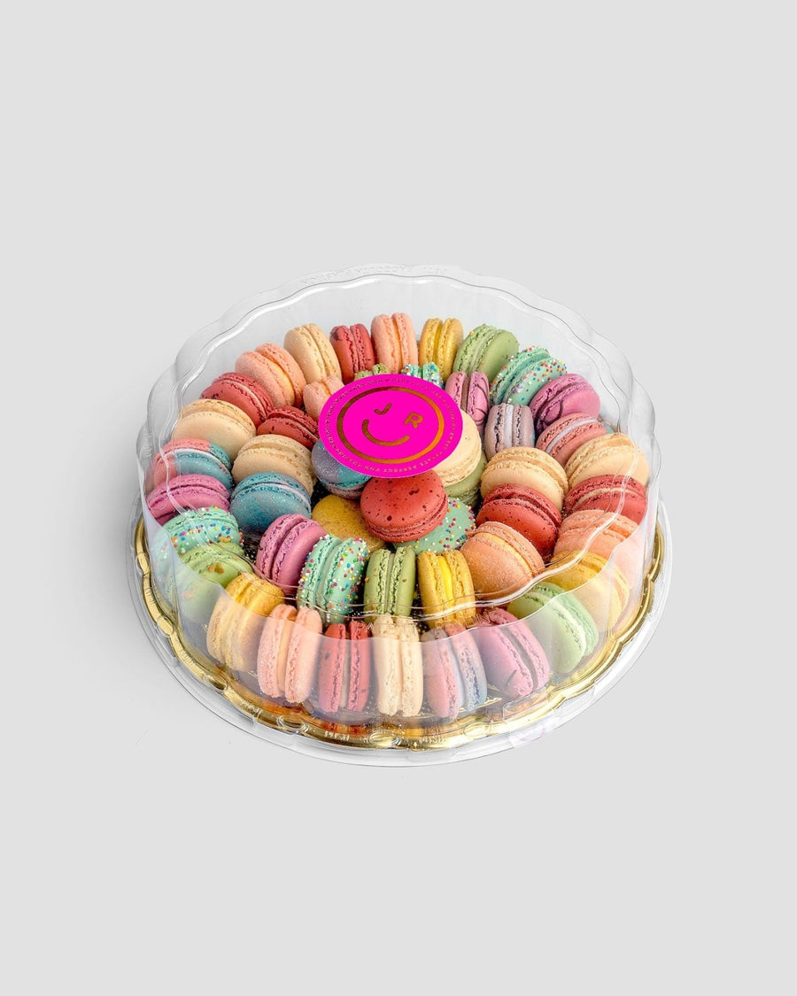 The Jenna Rae Cakes Macaron Party Platter features colorful macarons arranged in circles inside a round clear plastic container, topped with a pink smiley face decoration, set against a light gray background.