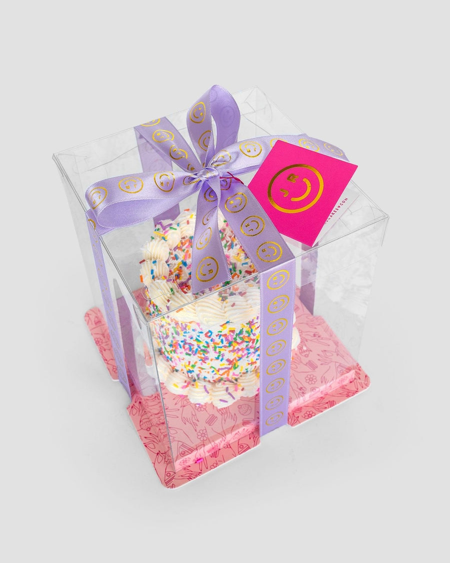 The Mini Vanilla Sprinkle Cake by Jenna Rae Cakes features white frosting with colorful sprinkles in a clear box, accented with a lavender ribbon of yellow smiley faces, pink smiley-face tag, and a pink patterned base.