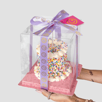 Someone holds the Jenna Rae Cakes Mini Vanilla Sprinkle Cake with white frosting and colorful sprinkles in a clear box tied with a purple smiley-face ribbon. The cake rests on a pink base and has a smiley-face tag attached.