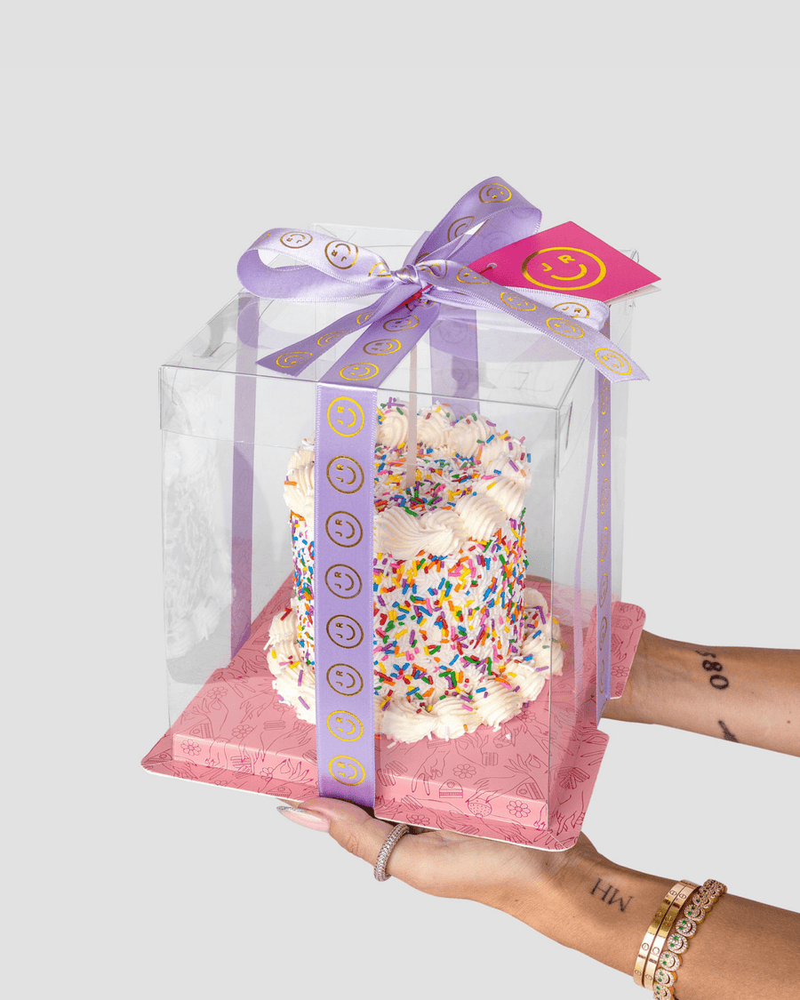 Someone holds the Jenna Rae Cakes Mini Vanilla Sprinkle Cake with white frosting and colorful sprinkles in a clear box tied with a purple smiley-face ribbon. The cake rests on a pink base and has a smiley-face tag attached.