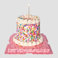 The Mini Vanilla Sprinkle Cake by Jenna Rae Cakes, topped with a single lit candle and covered in white frosting and colorful sprinkles, sits on a pink square board with whimsical line art against a plain gray background.