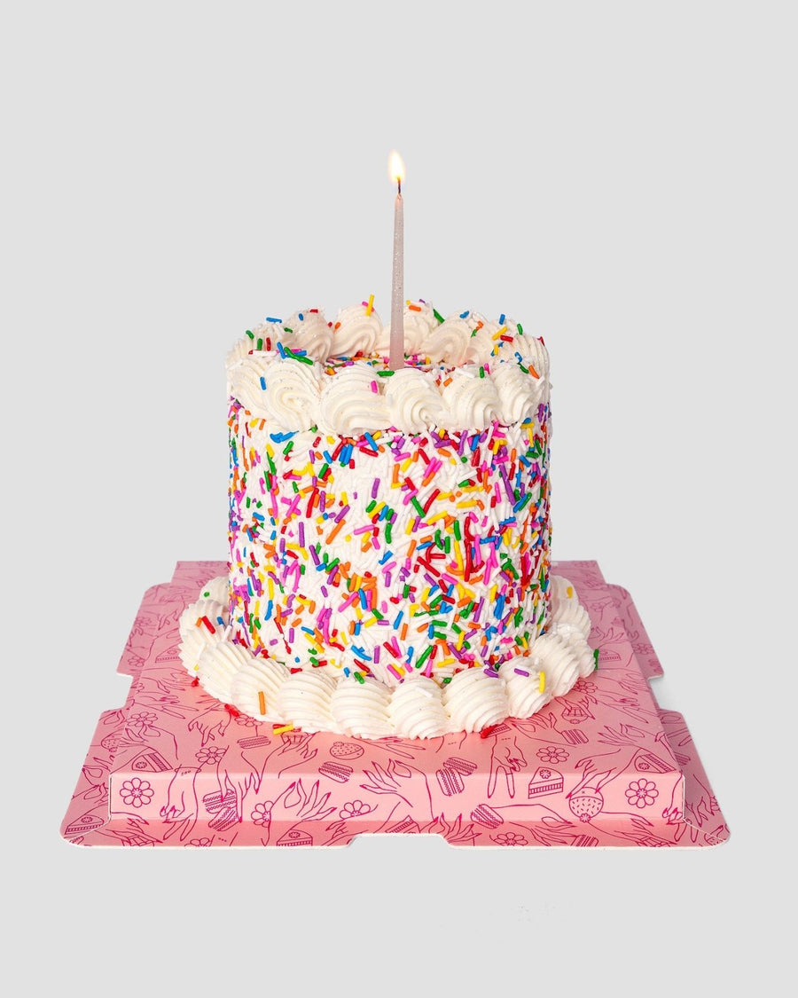 The Mini Vanilla Sprinkle Cake by Jenna Rae Cakes, topped with a single lit candle and covered in white frosting and colorful sprinkles, sits on a pink square board with whimsical line art against a plain gray background.