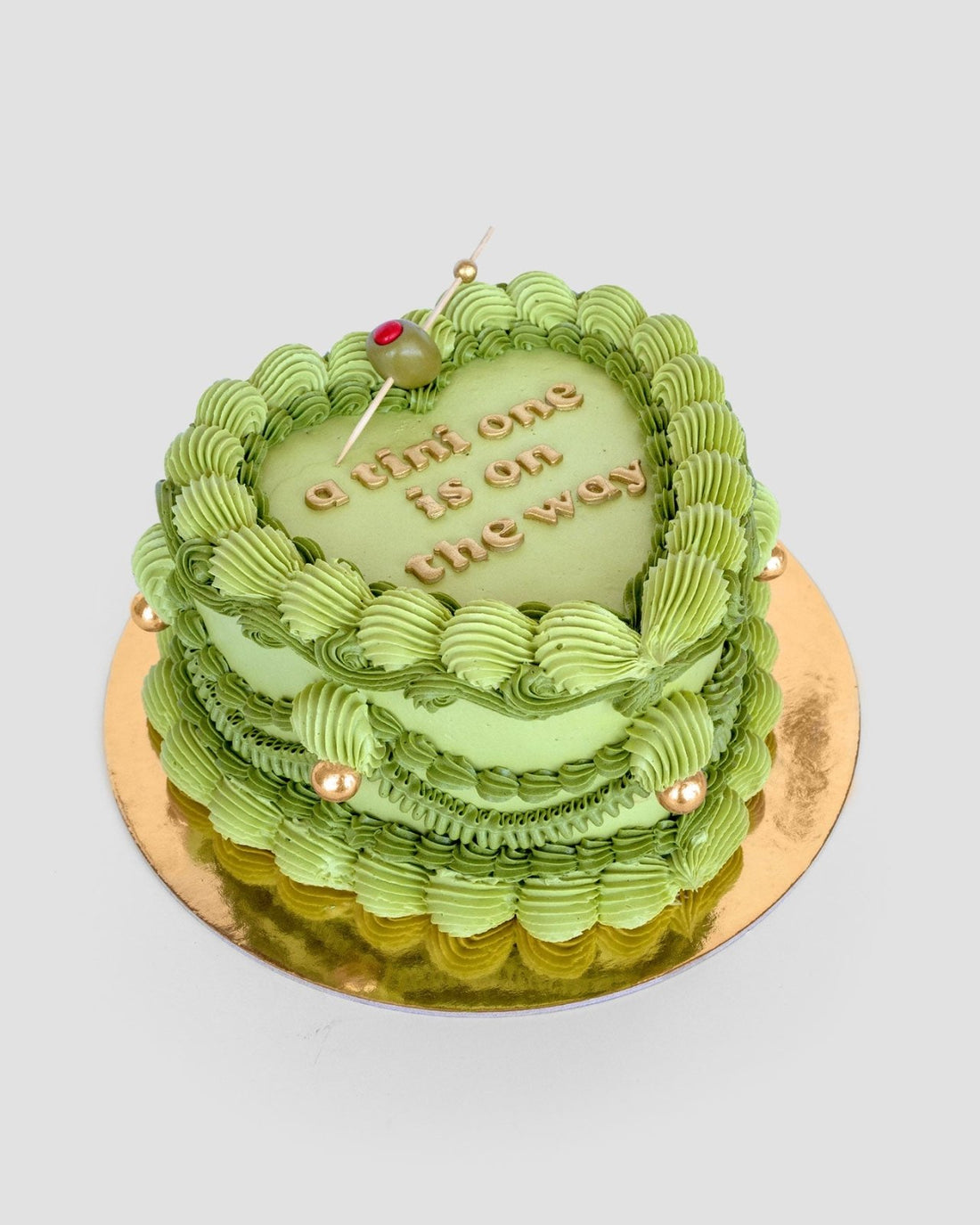 The Olive Martini Heart Cake by Jenna Rae Cakes features green icing, gold pearls, a cherry on top, and a little one is on the way in gold lettering. Shaped like a heart, this cake is presented on a gold board.