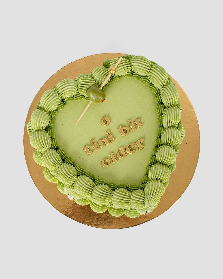 The Olive Martini Heart Cake by Jenna Rae Cakes is a heart-shaped green cake with piped frosting edges, topped with a green olive on a toothpick and a tini bit older written in gold letters, set on a gold cake board.