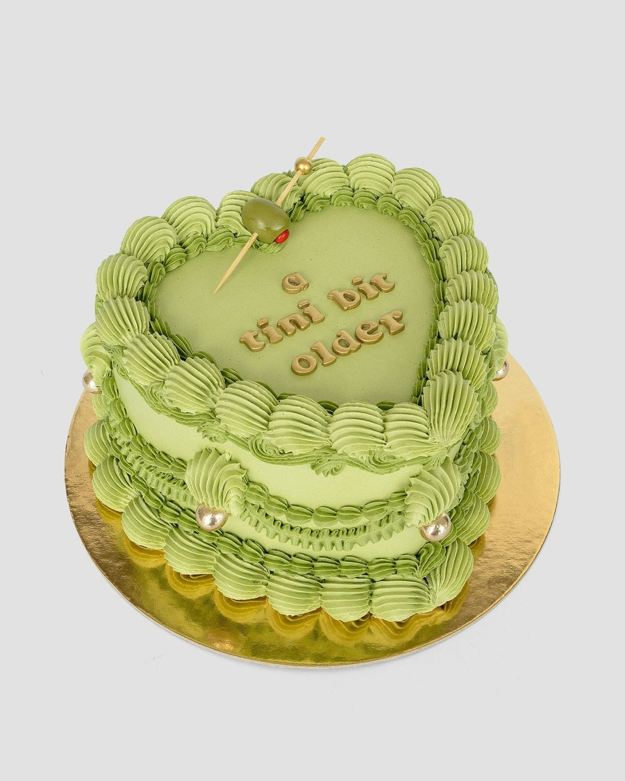 The Olive Martini Heart Cake by Jenna Rae Cakes features a heart-shaped green design with piped frosting, a green olive on a cocktail stick, golden pearls, and “a tini bit older” in gold letters atop a gold board.