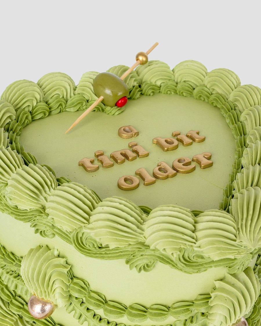 The Olive Martini Heart Cake by Jenna Rae Cakes features a heart-shaped green cake with piped frosting, gold a tini bit older lettering, and a cocktail olive on a toothpick.