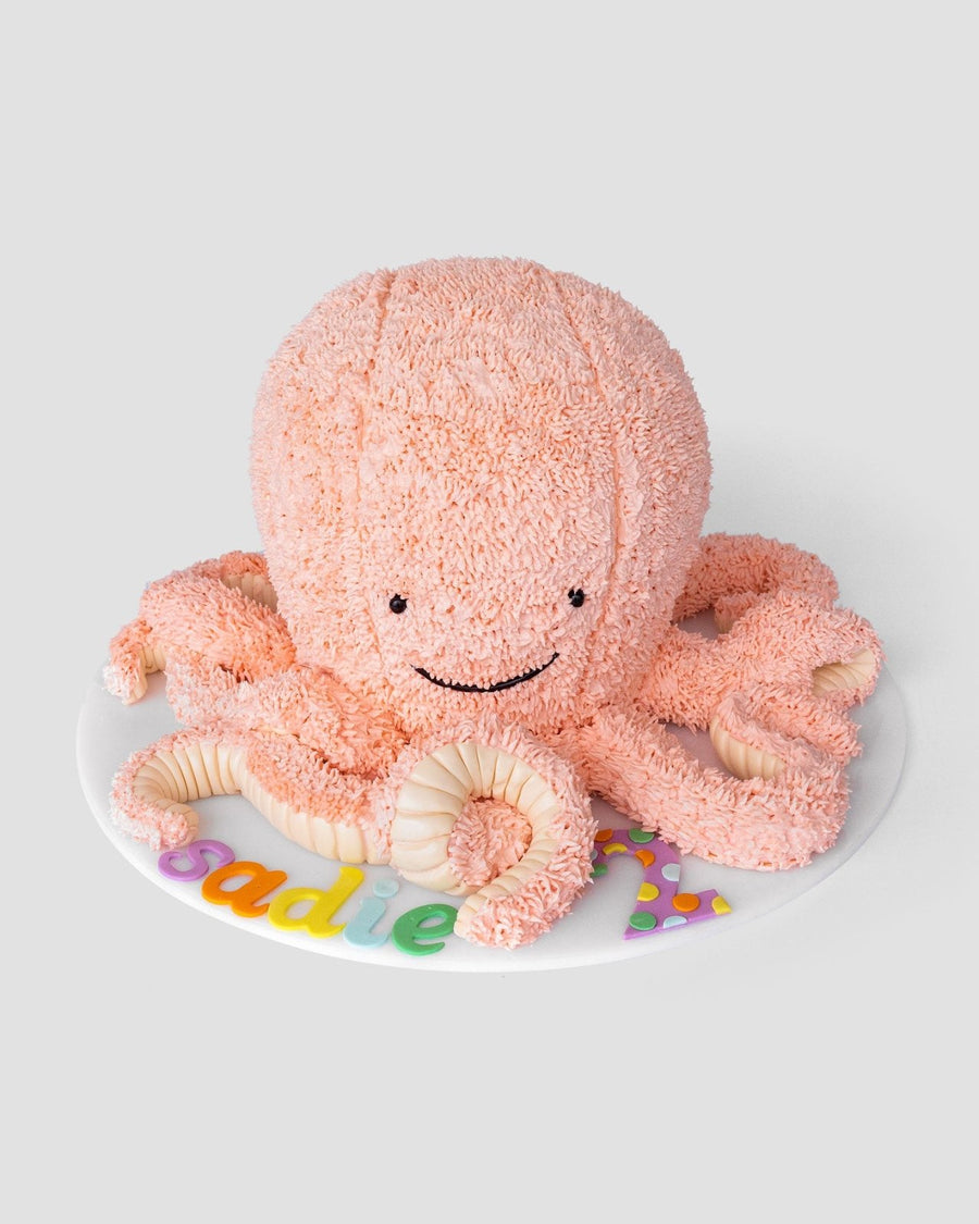 A pink, octopus-shaped cake with textured frosting, a smiling face, and curled tentacles sits on a white round board decorated with colorful letters.
