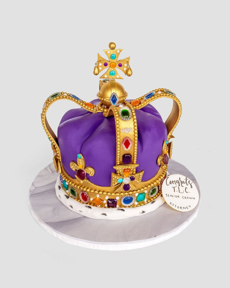 A One-of-a-Kind Cake by Jenna Rae Cakes shaped like a royal crown, adorned with gold accents and edible jewels on purple fondant, sits on a white board with a “Congrats MEC, Senior Crown Attorney” tag.