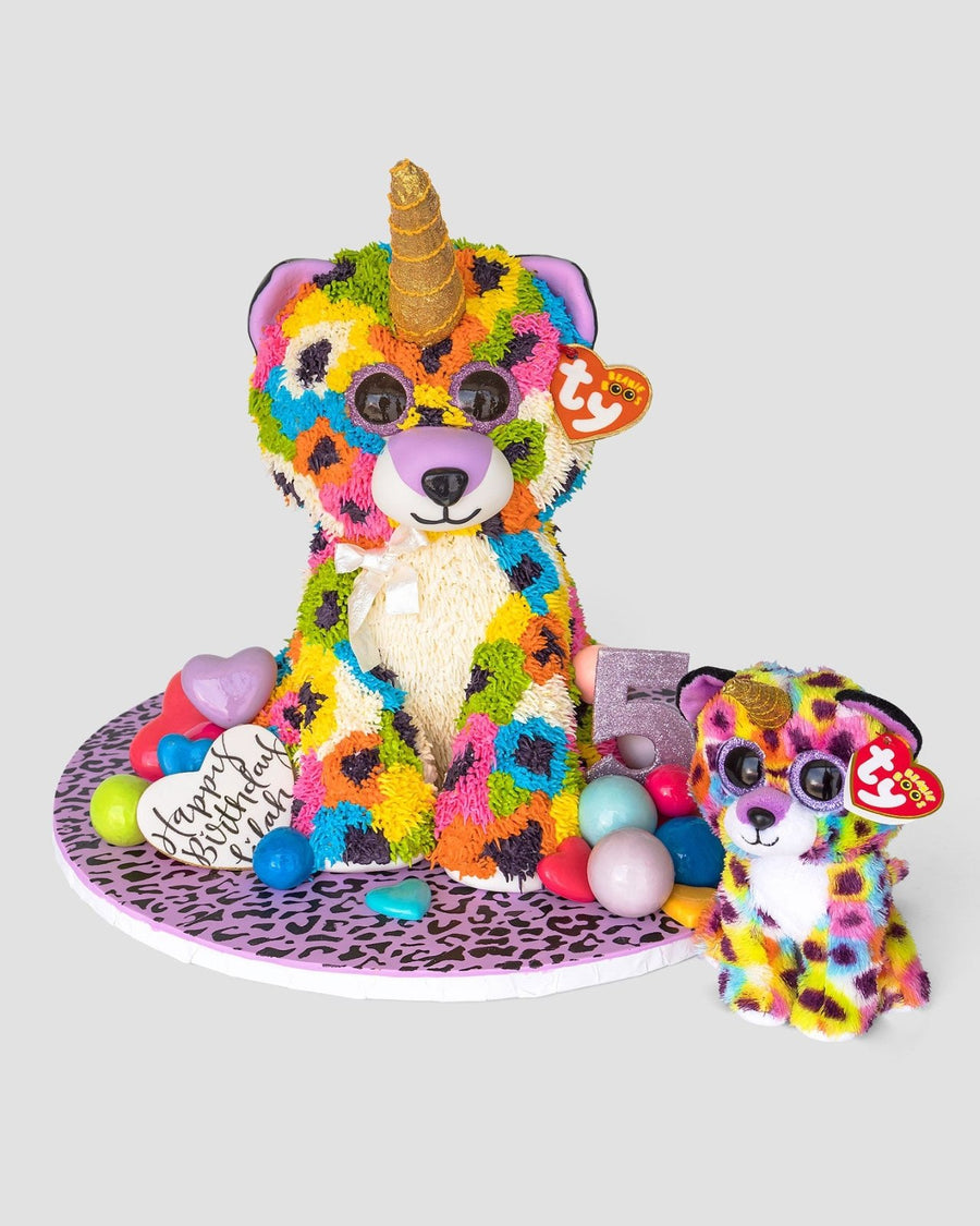 A vibrant unicorn bear cake from Jenna Rae Cakes’ One-of-a-Kind Cakes, featuring a gold horn, rainbow spots, and sunglasses, sits beside a matching Ty-tagged plush. The display includes balloons and a Happy Birthday message.