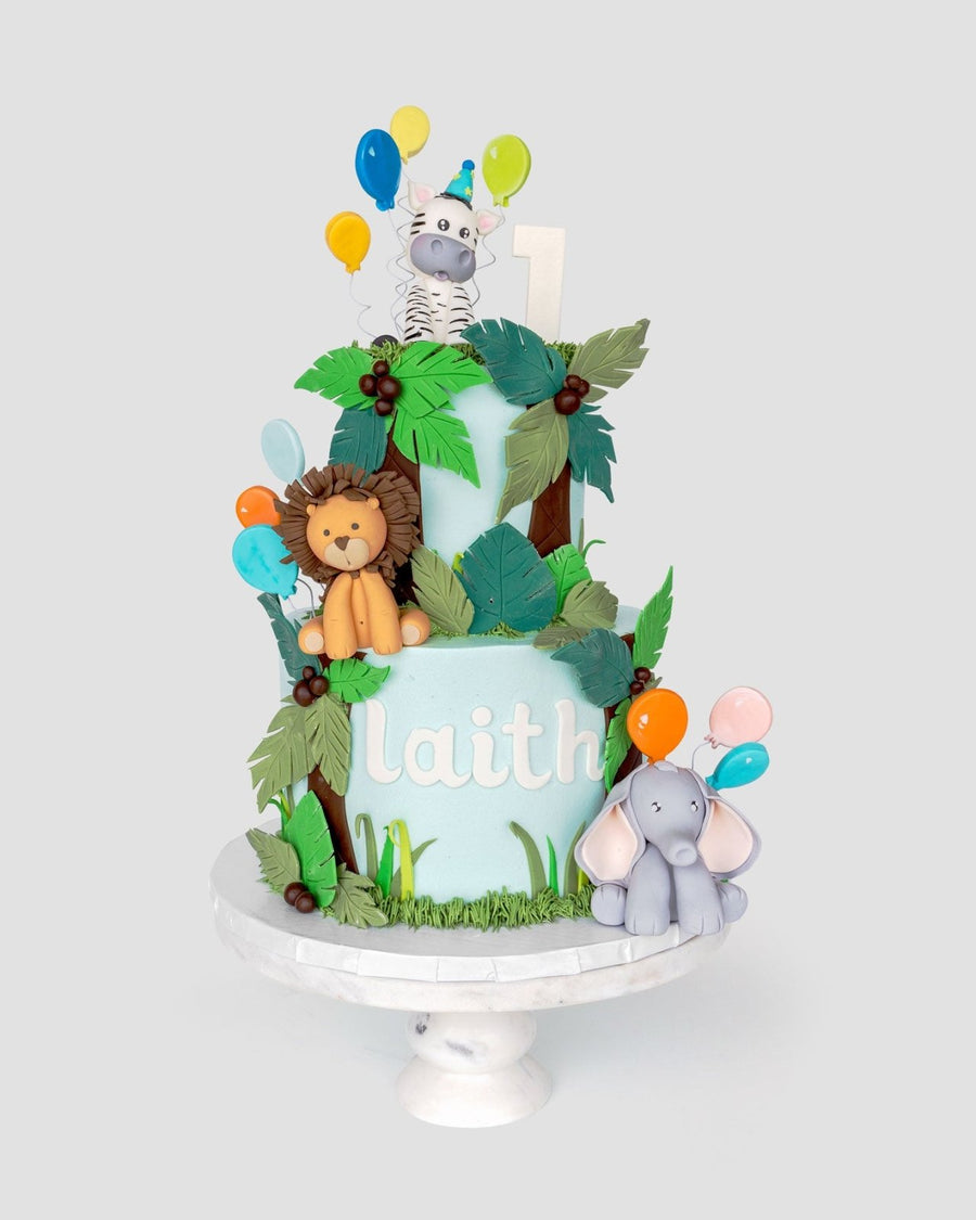 A two-tiered jungle-themed cake with fondant animals—a lion, zebra, and elephant—colorful balloons, green leaves, and the name laith on the side. The cake sits on a white stand with a number 1 topper.