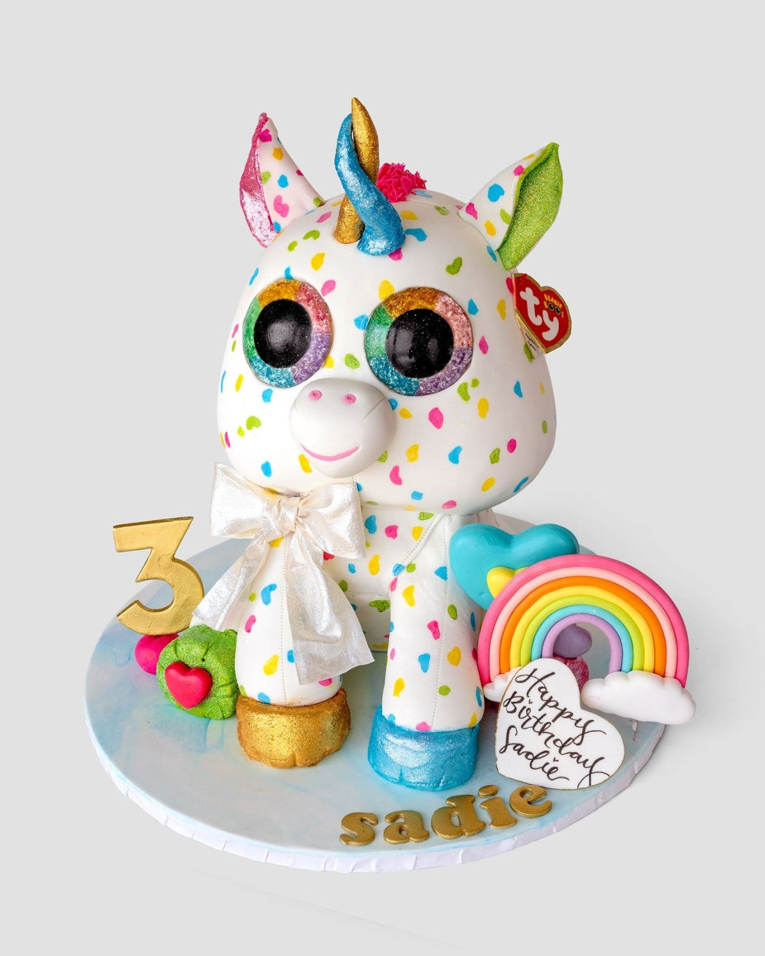 A colorful unicorn cake decorated with rainbow spots, large eyes, a gold horn, and ears. It has a gold “3,” a rainbow, a heart, and a “Happy Birthday Sadie” message on a white cake board.